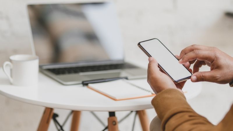 Hands holding a smartphone with a laptop computer in the background.