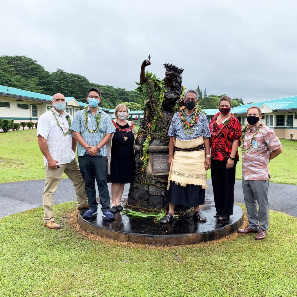 People standing next to a bronze statue. The statue is adorned with fresh lei.
