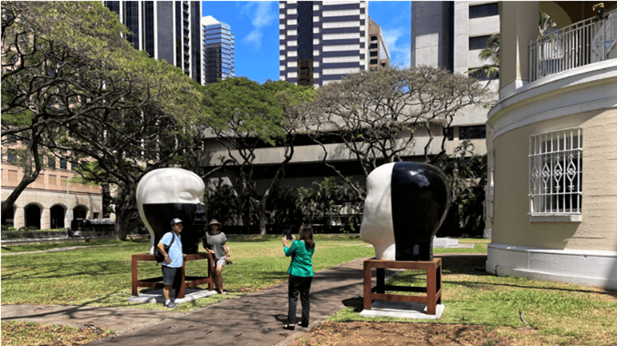 Two people posing for a photograph while a third person takes the photo in front of a large black and white sculpture of a human head. A second sculpture is on the othe rside of a walkway.