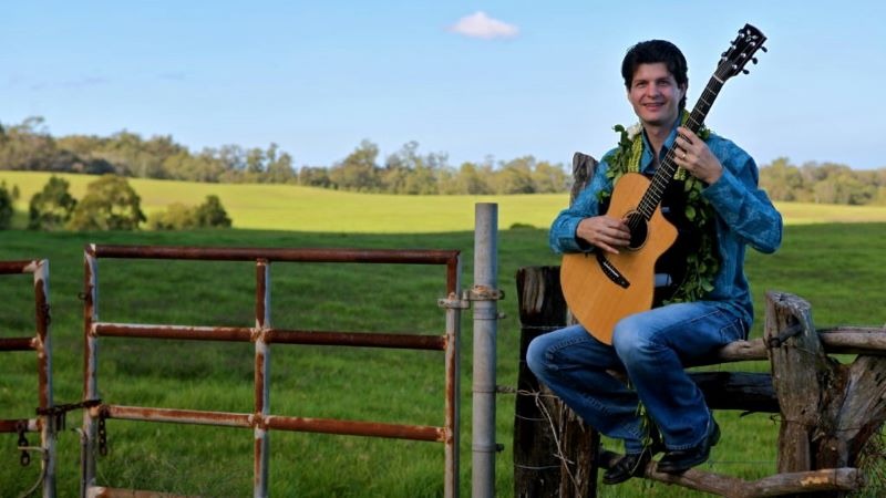 Jeff Peterson sitting on a fence and holding a guitar.