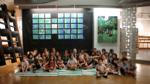 Preschool students sitting on mats in front of a display of their paintings.